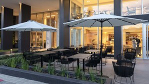 An outdoor seating area with tables and chairs, a coffee shop in the background, and a modern building facade. - Dots del Alto (Neuquén)