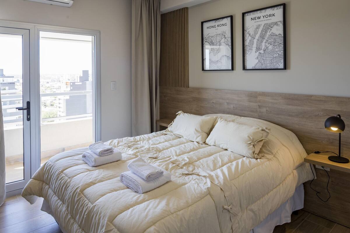 Cama bem arrumada com edredom branco, luminária de cabeceira e dois mapas emoldurados na parede.