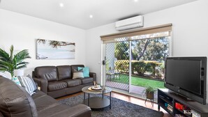 A modern living room with a leather sofa, a glass coffee table, a flat-screen TV, and a large sliding glass door leading to a garden. - Island Vibes Retreat on the Cowes Esplanade (Cowes)