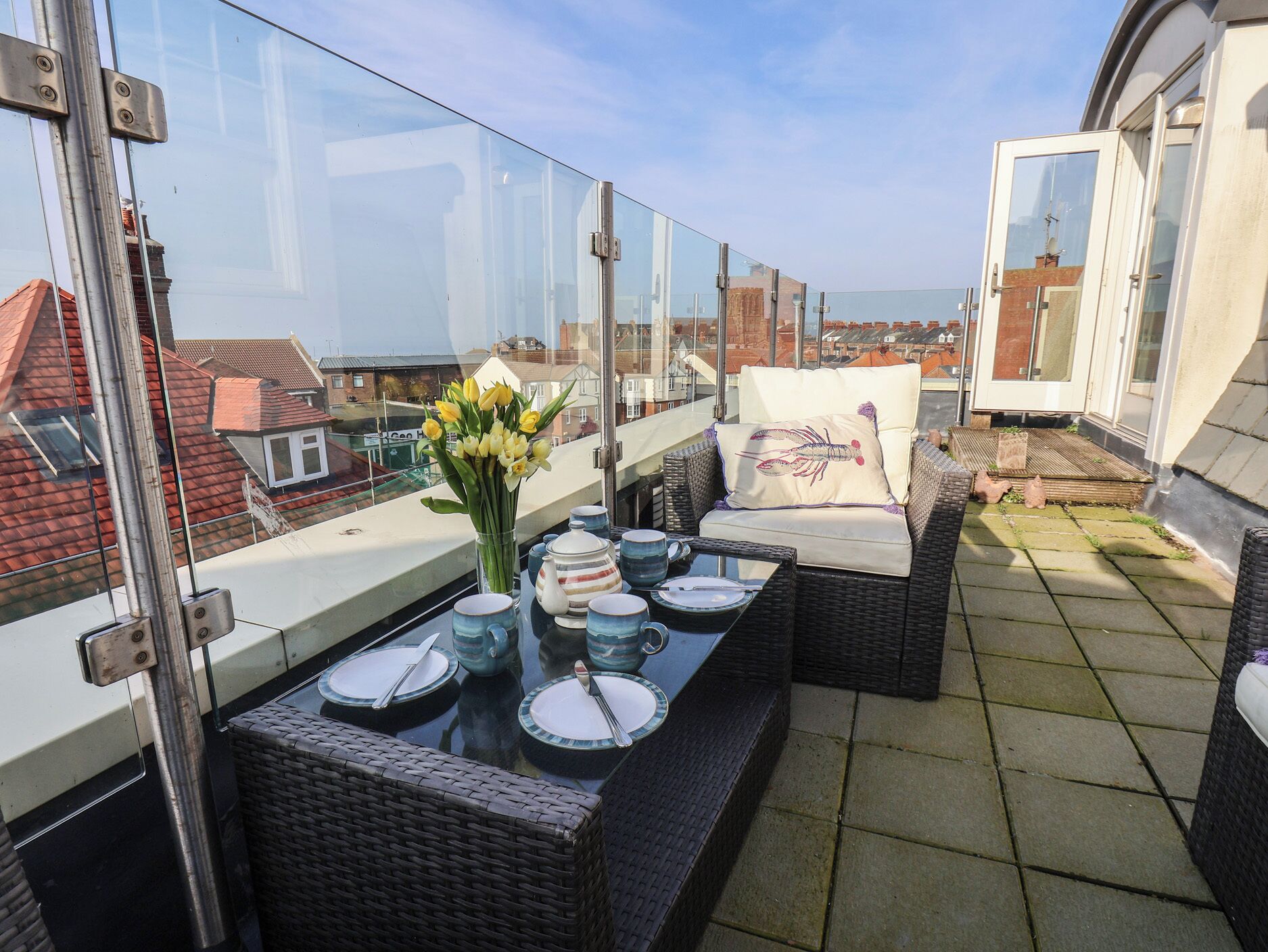 Ein Balkon mit Korbmöbeln, einem Glastisch mit Geschirr und Besteck sowie einer Vase mit gelben Blumen.