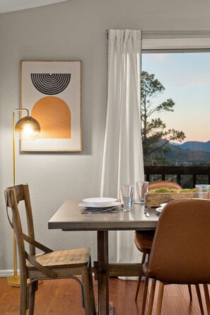 A dining area with a table set for two, a framed artwork above, and a view of a mountainous landscape through a window with white curtains. - Sedona`s Oceanfront Property • Spa, Views, Trails! (Sedona)