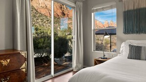 A bedroom with a large sliding glass door leading to a garden, a bed with white bedding, a nightstand with a lamp, and a dresser. - Sedona`s Oceanfront Property • Spa, Views, Trails! (Sedona)