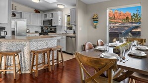 A modern kitchen with white cabinets, a central island with stools, and a dining area with a table set for four. - Sedona`s Oceanfront Property • Spa, Views, Trails! (Sedona)