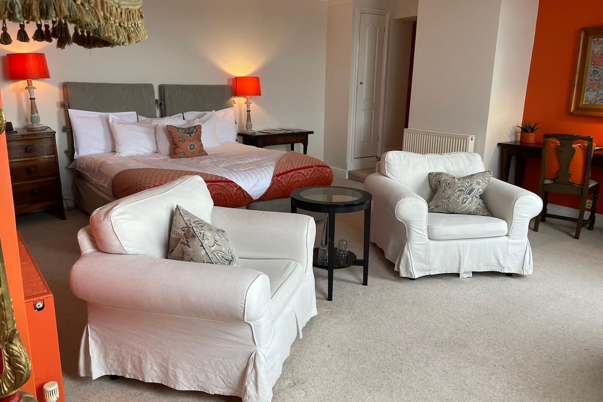 A hotel room with a bed, two white armchairs, a small table, and a red lamp.