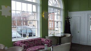 A room with a green wall, a patterned sofa, two chairs, a small table, and large windows with a view of buildings and parked cars outside.