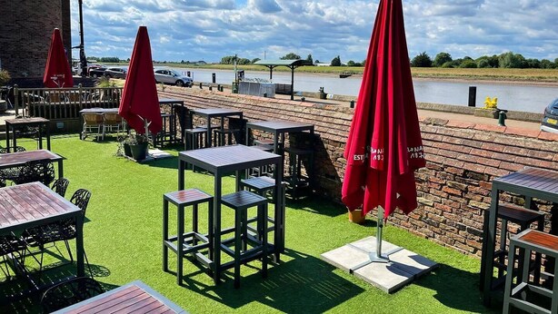 An outdoor seating area with tables and chairs, red umbrellas, and a view of a river.