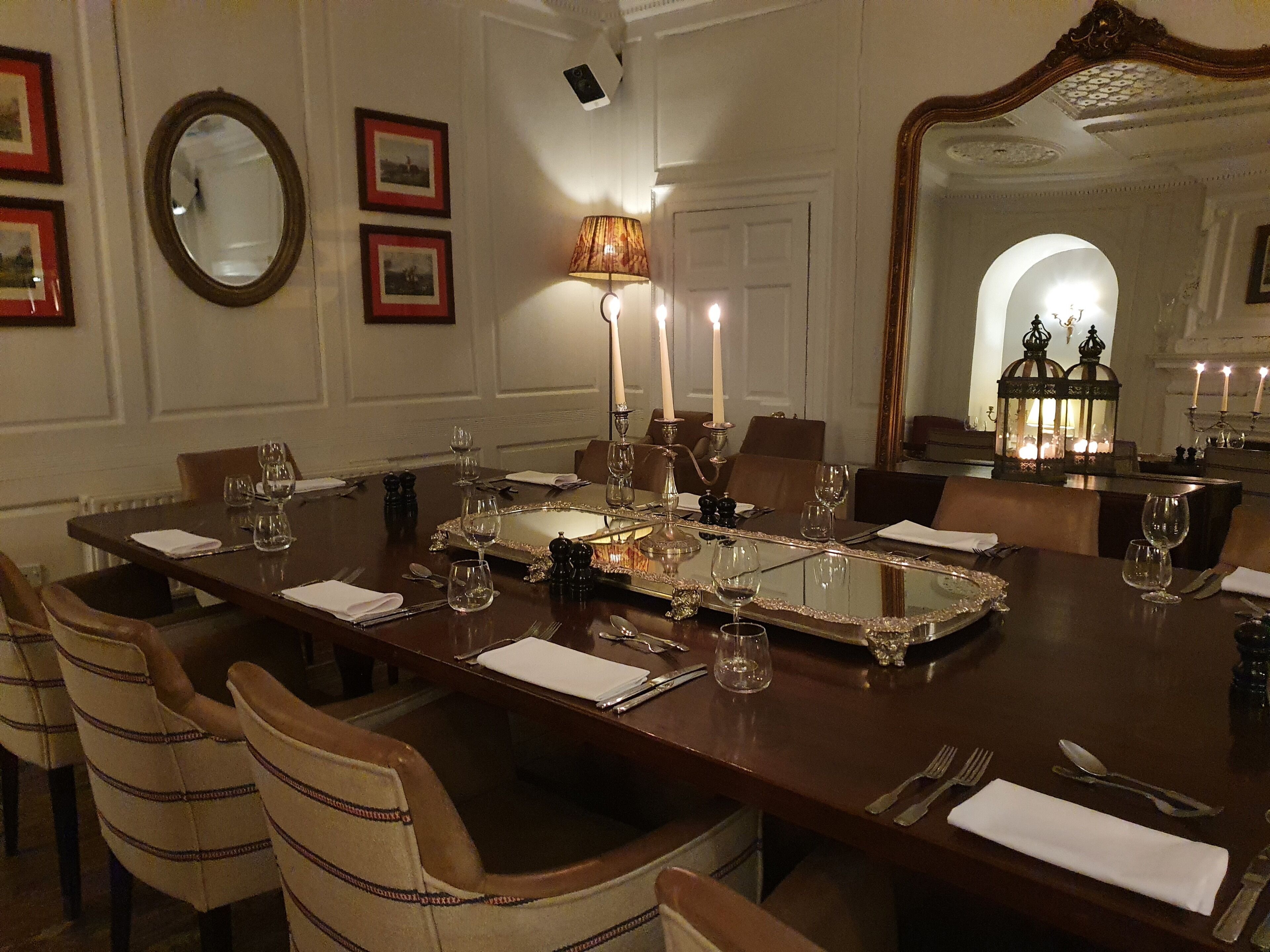 Un comedor elegante con una larga mesa dispuesta para varios invitados, que incluye un espejo, cuadros enmarcados y una araña.