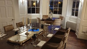 A formal dining room with a long table set for a meal, flanked by chairs, and large windows with curtains.