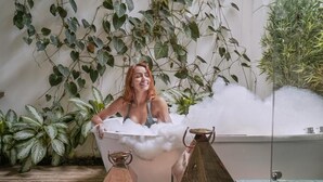 Una persona relajándose en una bañera llena de burbujas, rodeada de exuberante follaje y muebles rústicos.