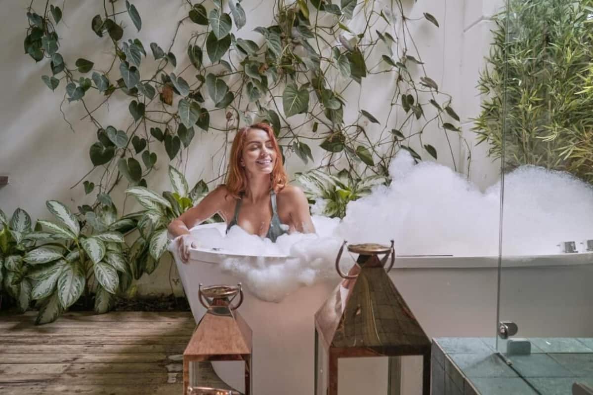 Una persona relajándose en una bañera llena de burbujas, rodeada de exuberante follaje y muebles rústicos.