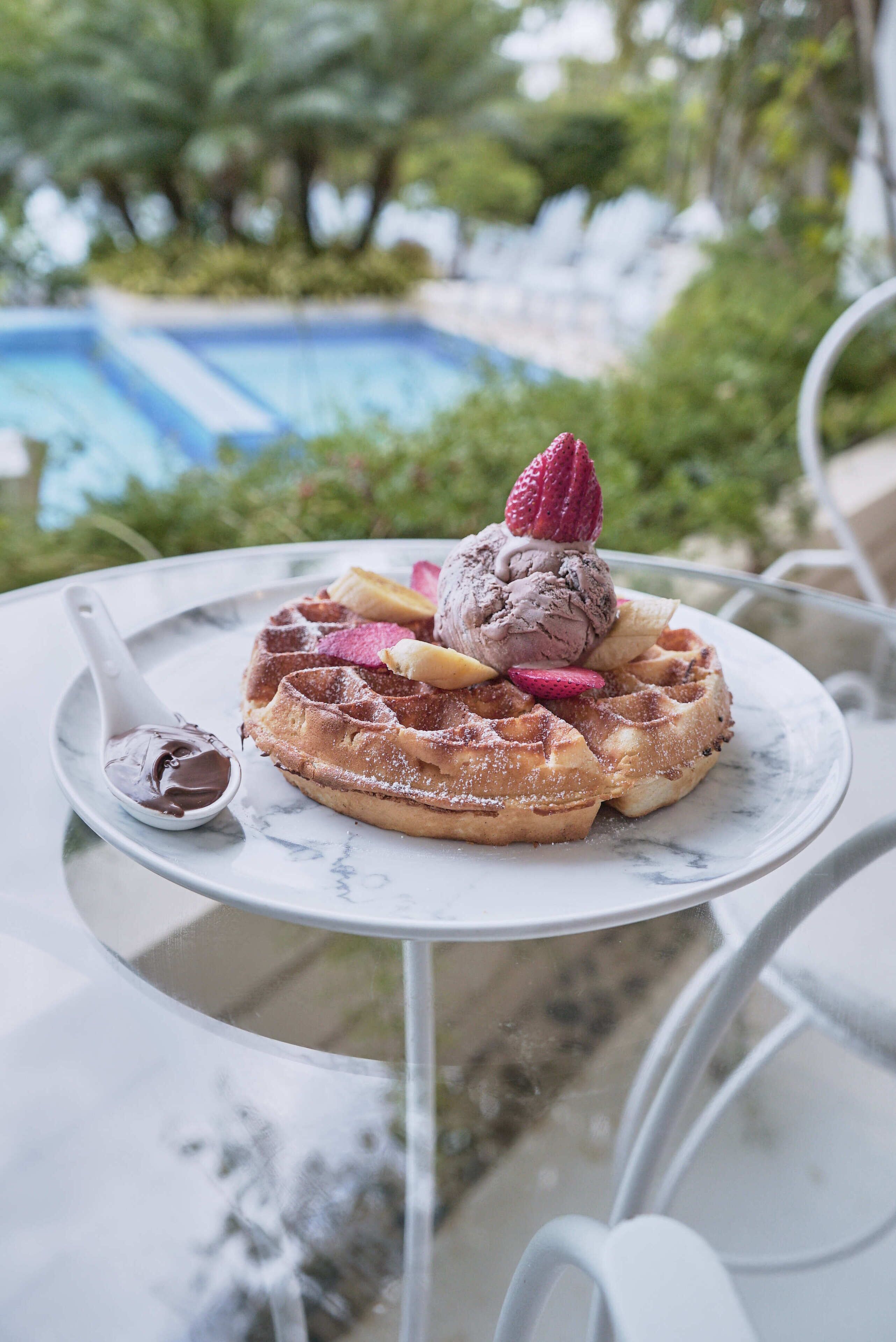 A waffle with ice cream and fruit on a glass table near a pool.