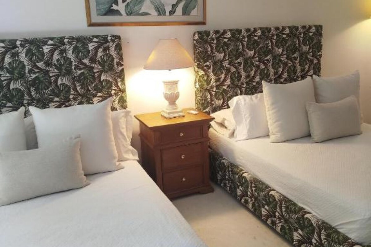 Una habitación de hotel con dos camas, una mesita de noche con lámpara y un cuadro con motivos tropicales en la pared.