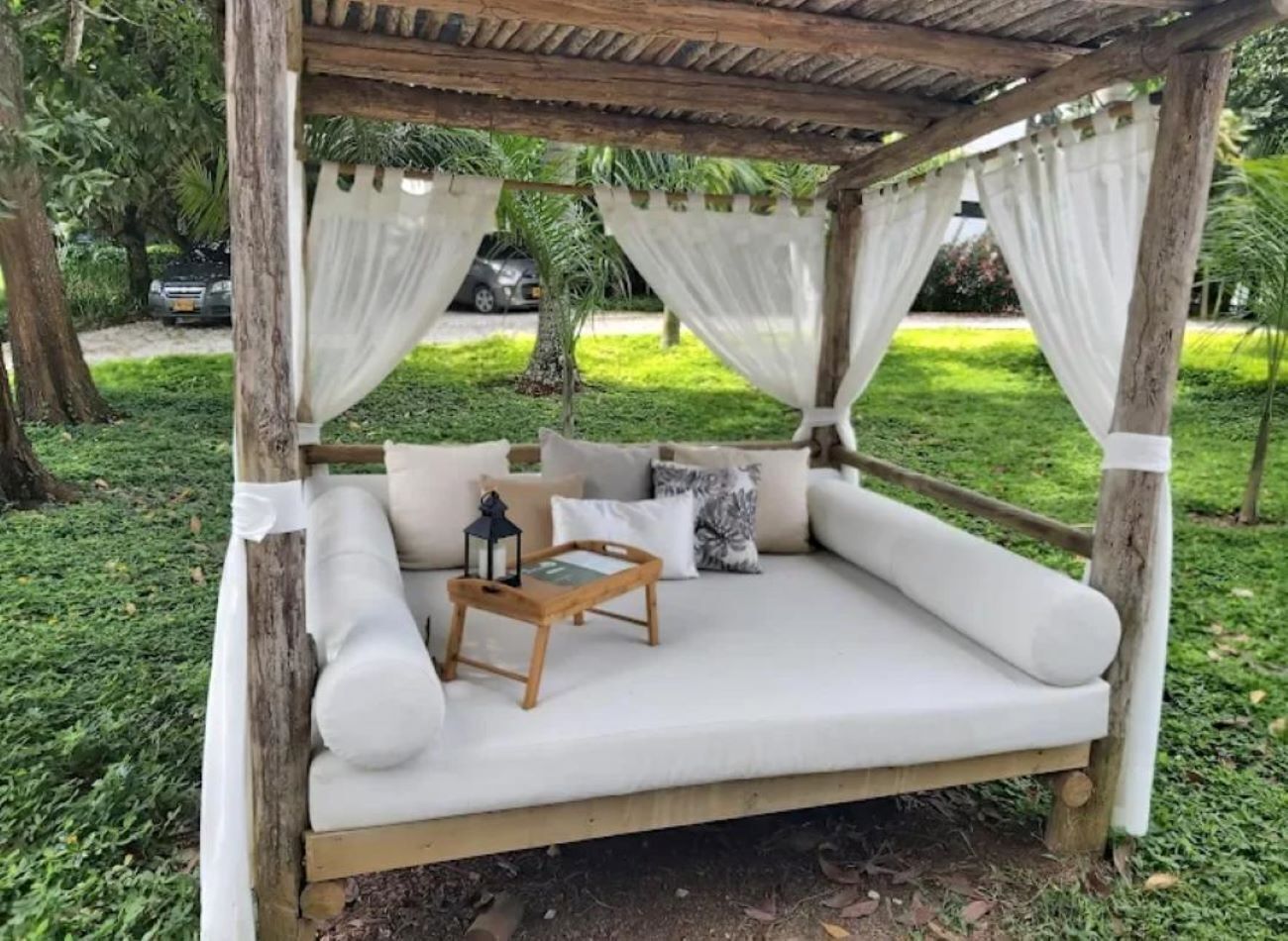 A wooden swing bed with white cushions, a small wooden table with a lantern, and white curtains.