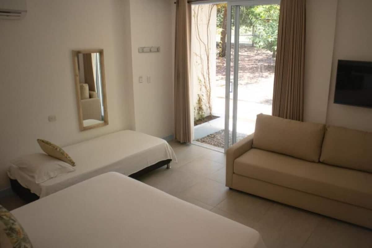 Habitación de hotel con dos camas, un sofá, una puerta corrediza de vidrio que da a un espacio exterior y un televisor montado.