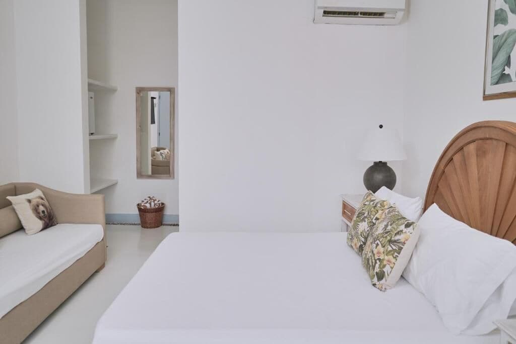 A bedroom with a bed, a bedside table with a lamp, a mirror, a small plant, and a sofa.