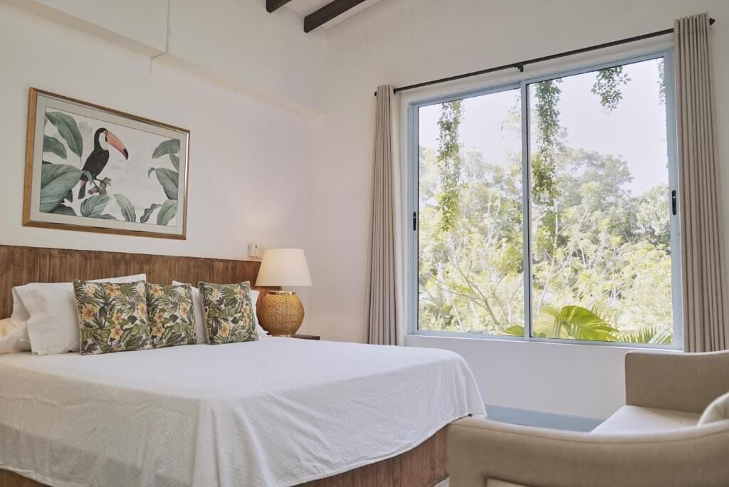 A bedroom with a bed, a chair, a window with curtains, a lamp, and a framed picture on the wall.