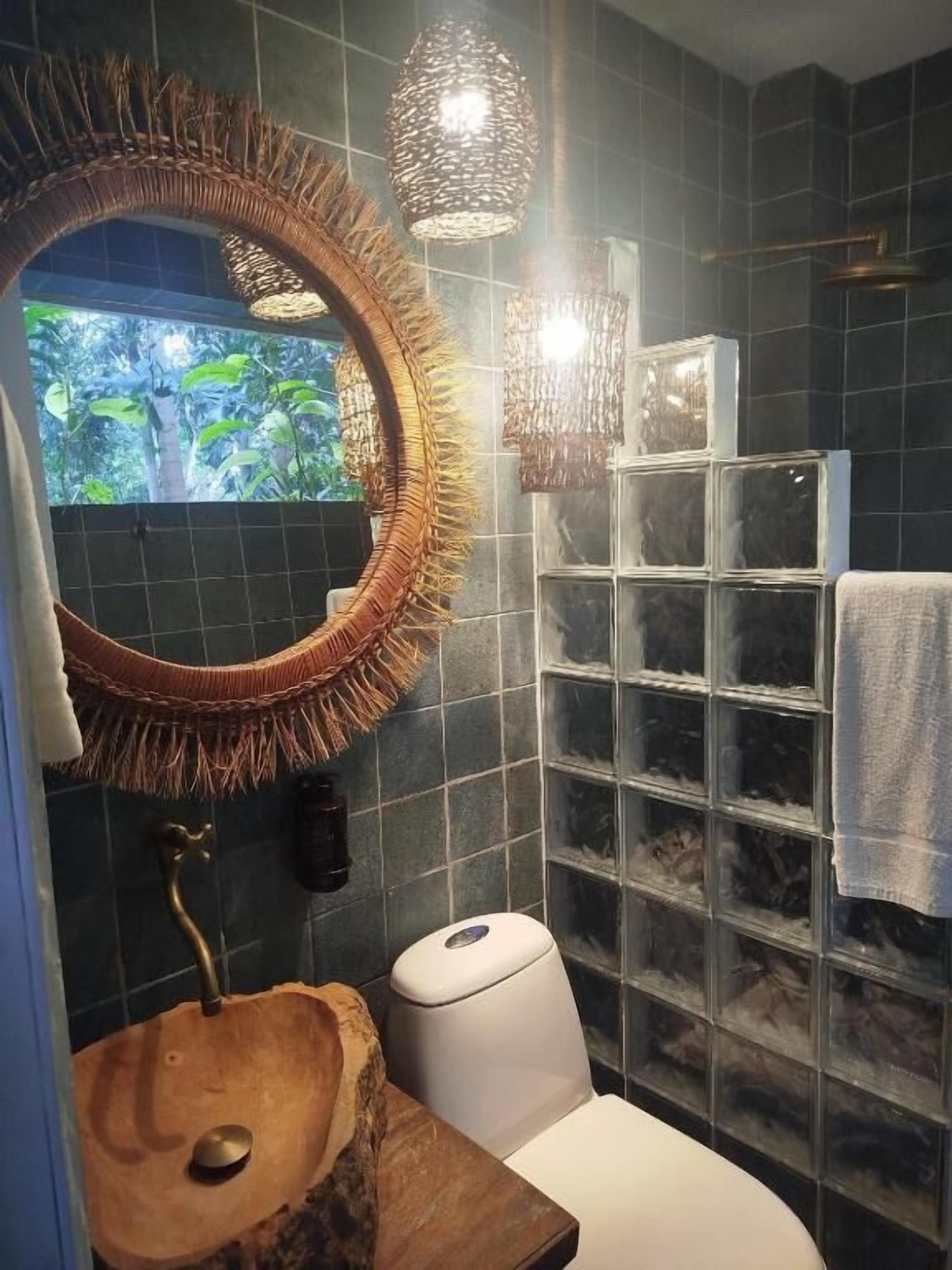 A bathroom with a wooden sink, a round mirror, a toilet, and a window with a view of greenery.