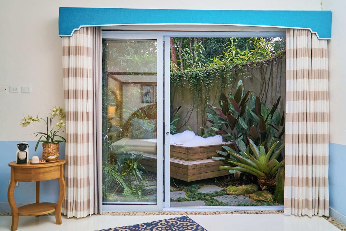 Una habitación con puerta corrediza de vidrio que da a un jardín, una pequeña mesa de madera con una planta y un banco con cojín.