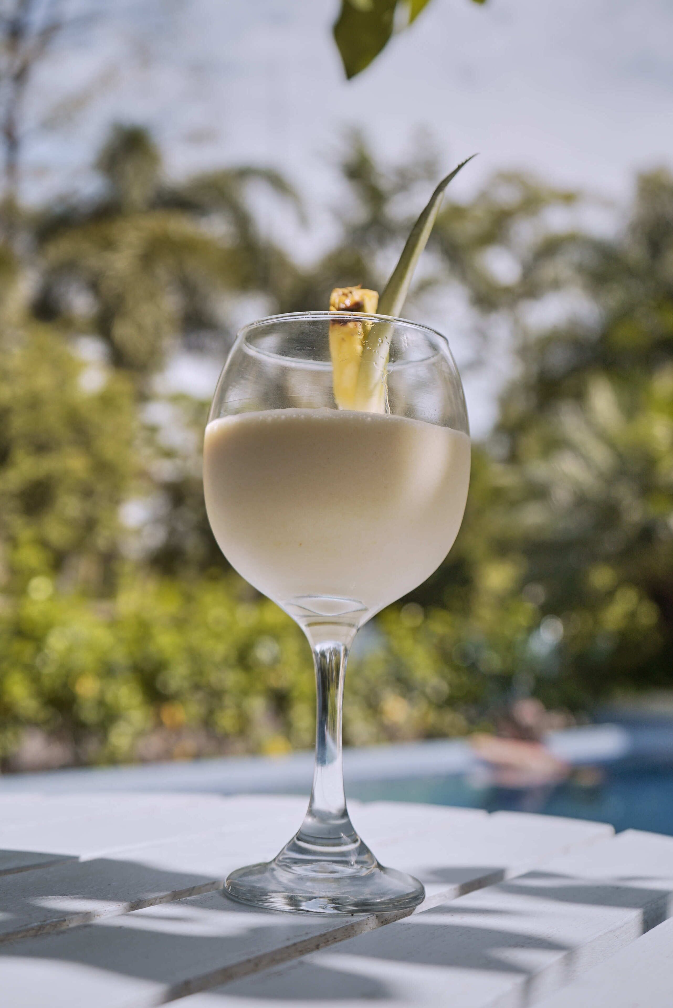 Un vaso transparente con una bebida cremosa, un adorno y una pajita, sobre una superficie blanca con una piscina y follaje al fondo.