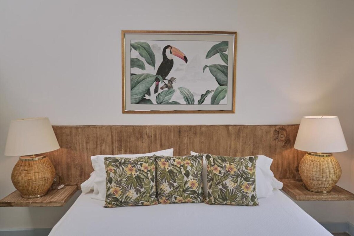 Habitación de hotel con una cama, dos lámparas de noche, un cuadro enmarcado de un ave tropical y un cabecero de madera.
