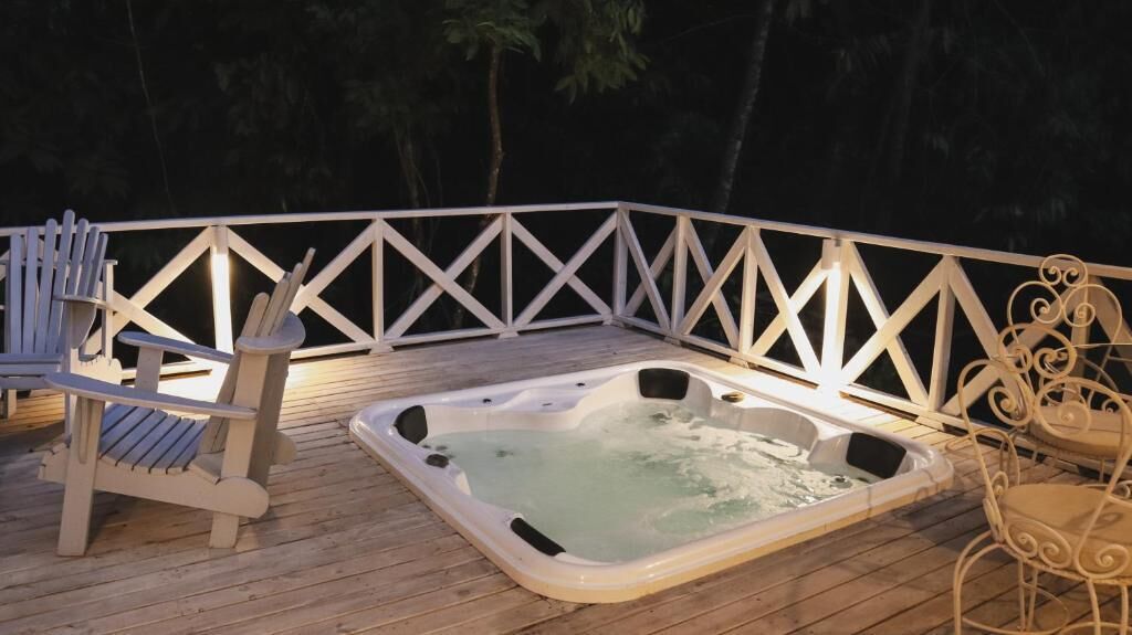 A wooden deck with a hot tub, Adirondack chairs, and a white railing.