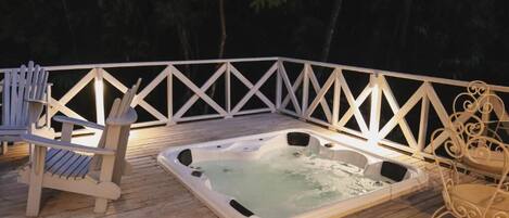 A wooden deck with a hot tub, Adirondack chairs, and a white railing.