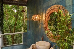 A bathroom with a large mirror, a wooden basin, and a view of lush greenery.