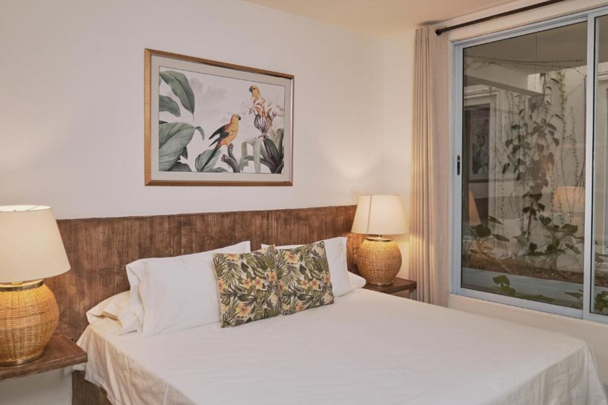 Ein Hotelzimmer mit einem Bett, zwei Nachttischlampen, einem gerahmten Bild an der Wand und Blick ins Grüne durch ein Fenster.