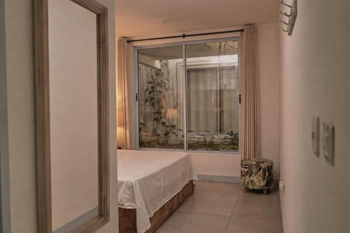 Un pasillo estrecho con una cama, un ventanal con cortinas y un taburete pequeño.