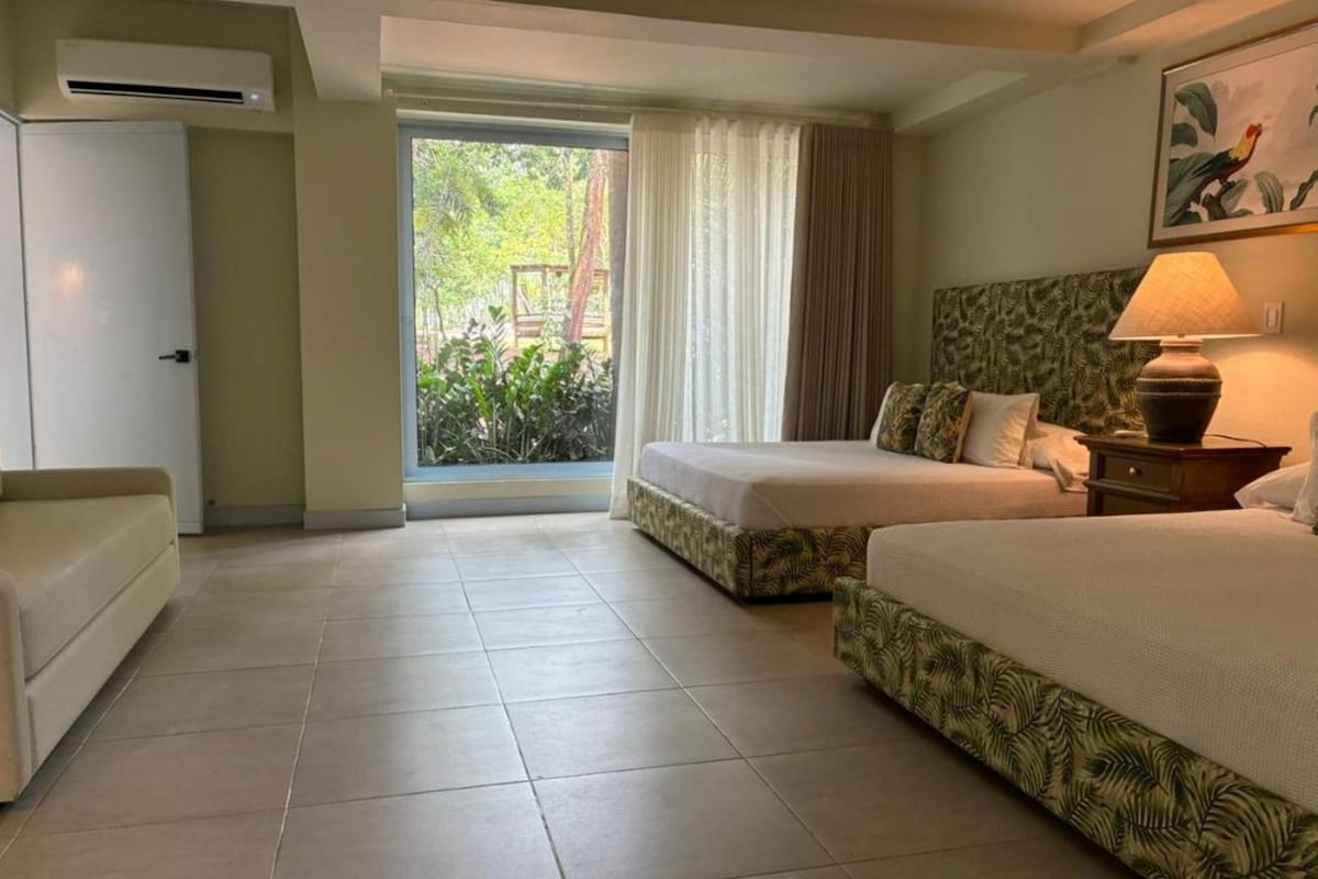Habitació d'hotel amb dos llits, un sofà, una finestra amb cortines i un quadre al mur.