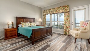 A bedroom with a wooden bed, a nightstand, a chair, and a window with curtains. - Screamin' Peace 1614 Cadillac 3 Bedroom Home by RedAwning (Dauphin Island)
