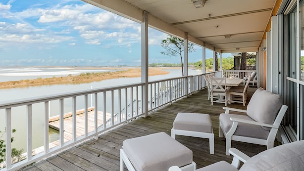 A covered outdoor area with a wooden deck, white cushioned chairs, and a view of a river and distant landscape. - Screamin' Peace 1614 Cadillac 3 Bedroom Home by RedAwning (Dauphin Island)