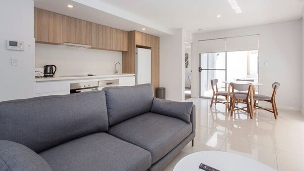 A modern living room with a grey sofa, a white coffee table, and a dining area with wooden chairs and a white table. - Stirling Apartment 1- Modern Central Apartment (Yokine)