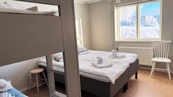 Een hotelkamer met een stapelbed, een eenpersoonsbed, een raam met uitzicht op een besneeerd landschap, een radiator en een witte stoel.