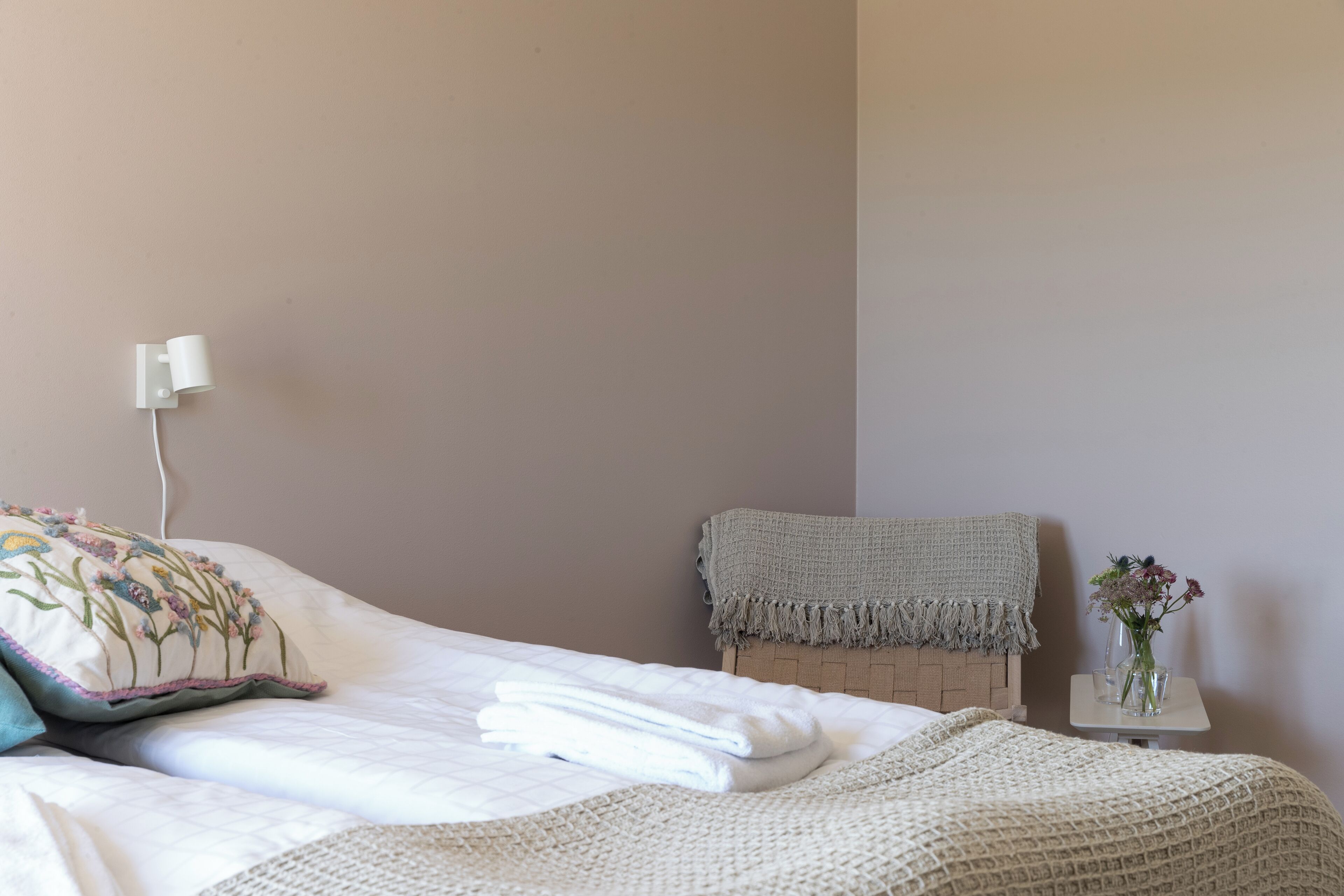 Un letto rifatto con asciugamani piegati, un cuscino decorativo, un comodino con un vaso di fiori e una piccola sedia con una coperta.