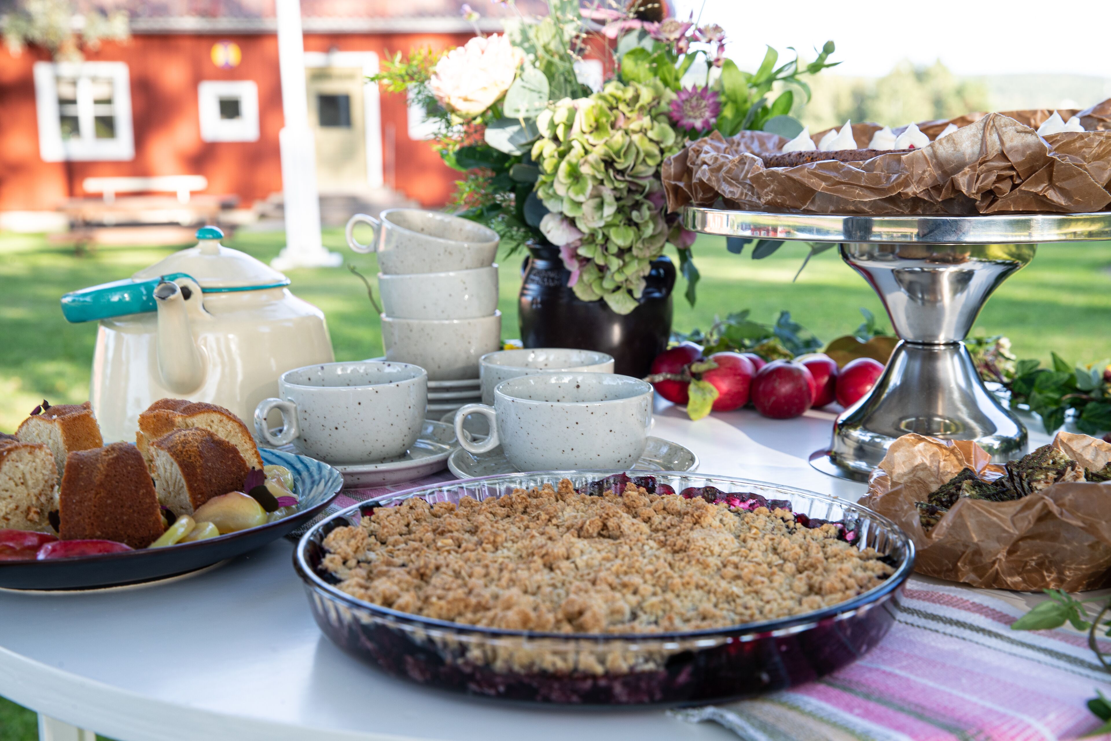 Ein Tisch mit verschiedenen Speisen, darunter ein Kuchen, Brot und eine Schüssel mit Streuseln, was auf ein Picknick oder eine Mahlzeit im Freien hindeutet.