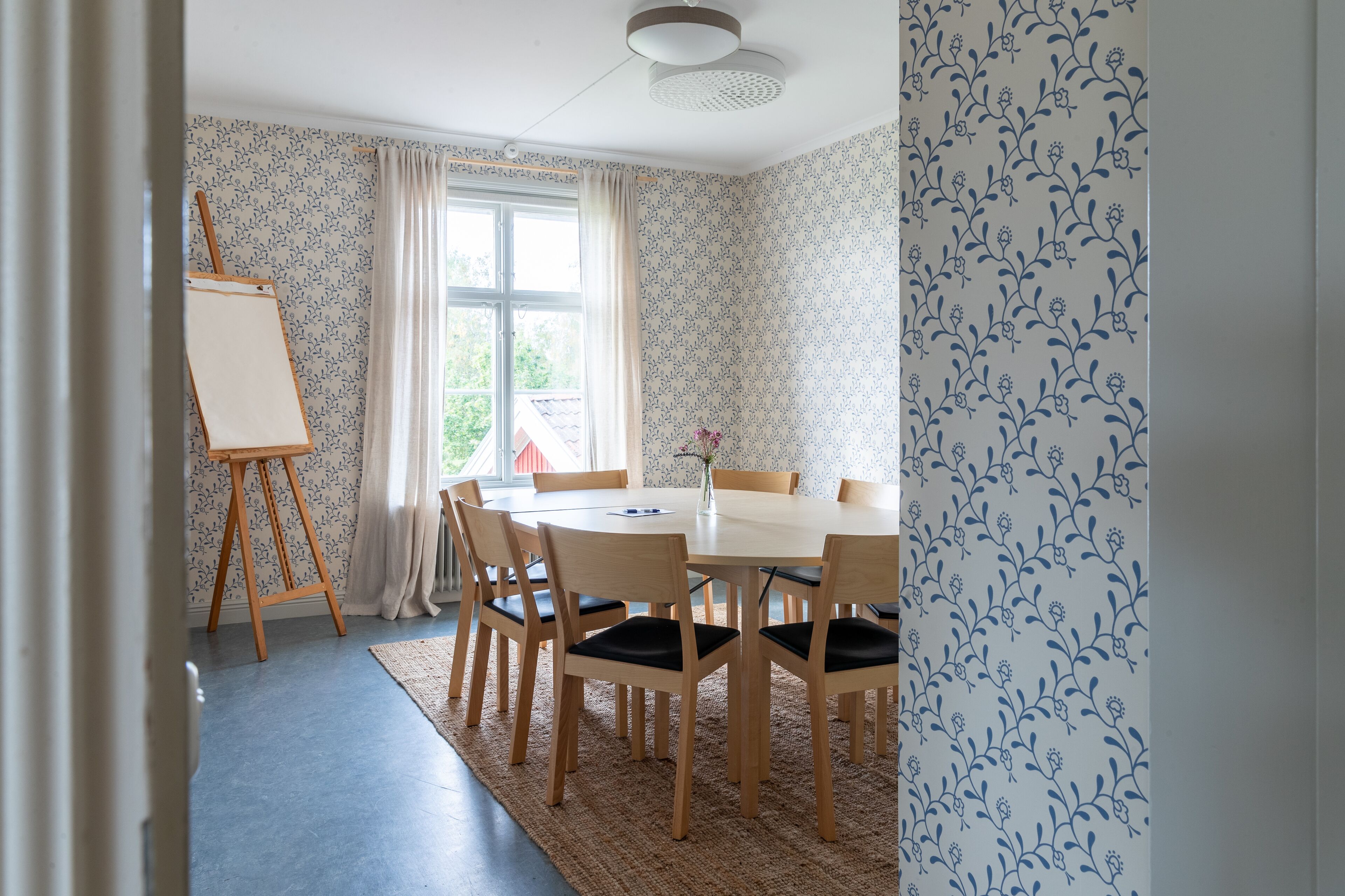 A dining room with a round table, six wooden chairs, a small easel, and a window with curtains.