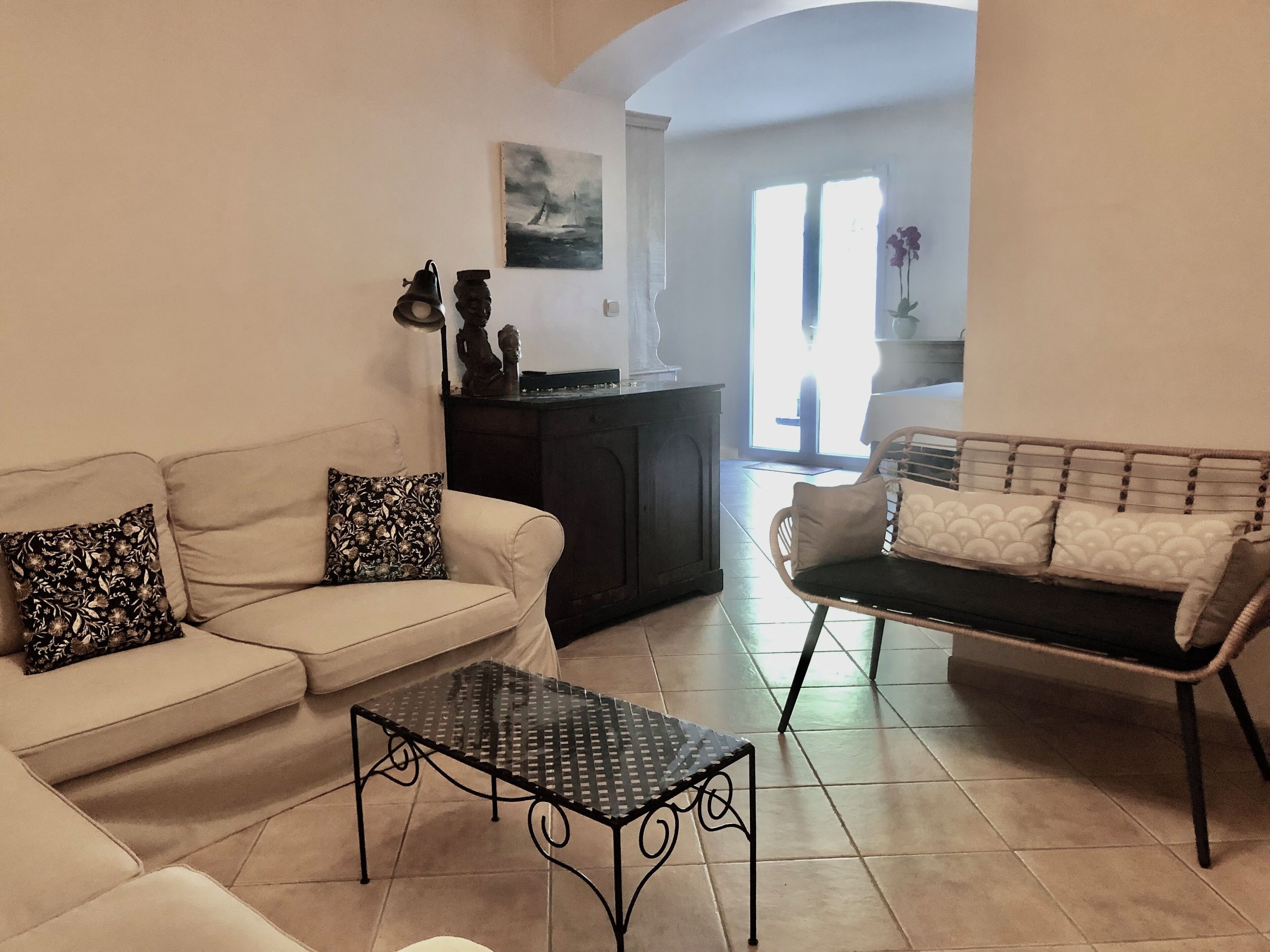 A living room with a sofa, a coffee table, and a wicker chair. There is a cabinet with decorative items and a painting on the wall.