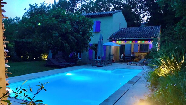 A swimming pool with a tiled deck, surrounded by greenery and a building with outdoor seating.