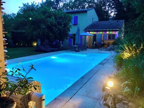 Piscina com deck ladrilhado, rodeada por vegetação e um edifício com área de estar externa.