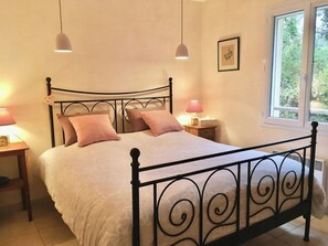 Quarto com estrutura de cama em metal, duas luminárias de apoio, janela com vista para área verde e um quadro na parede.