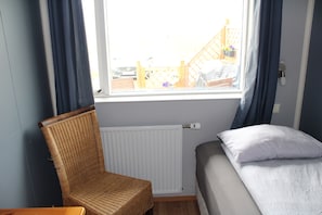 A room with a bed, a wicker chair, a window with curtains, and a white radiator.