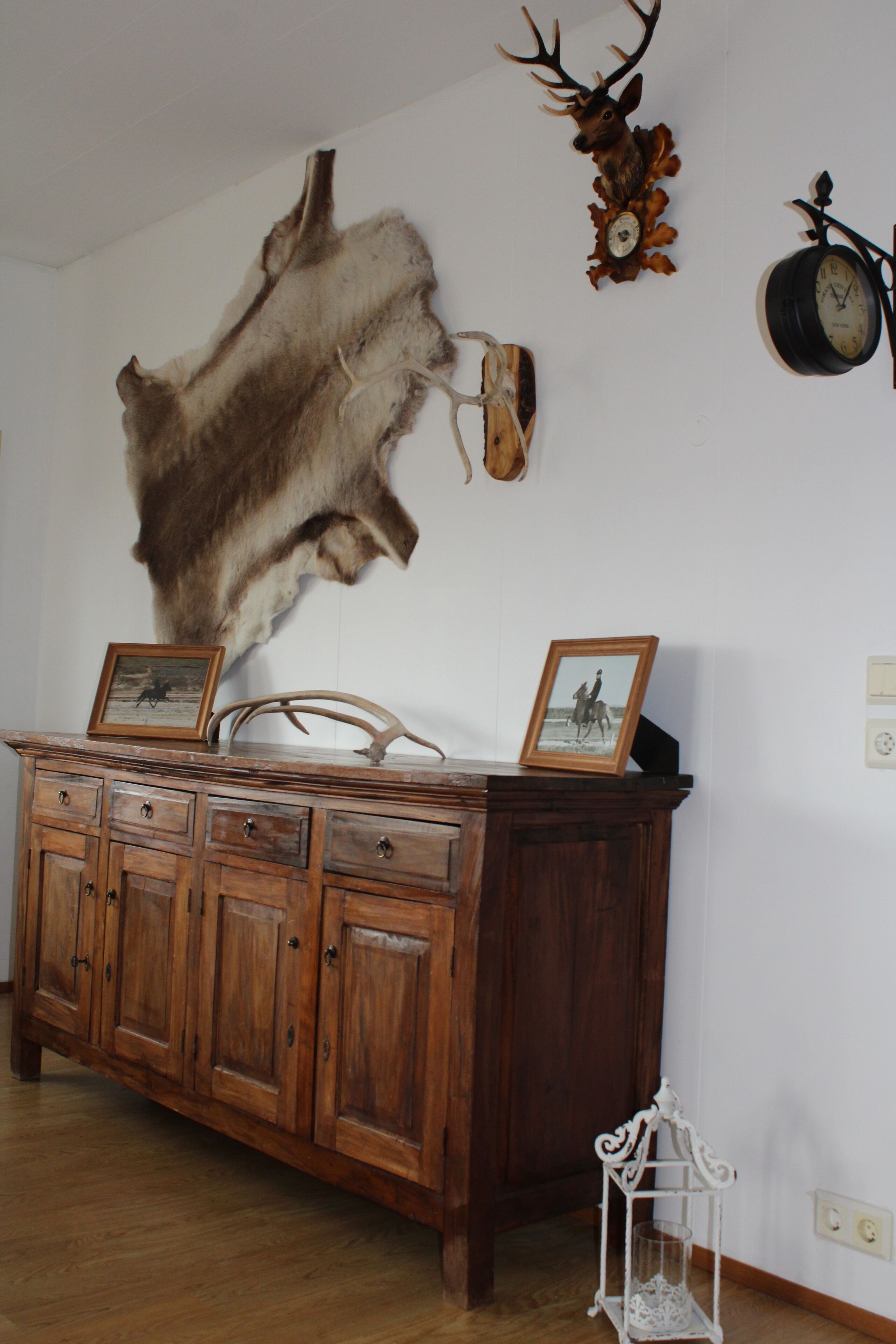 Una habitación con un tocador de madera, una cabeza de ciervo montada, un reloj y un cuadro en el tocador.