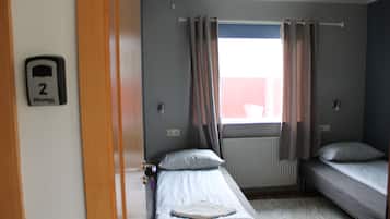 A small, single-room hotel room with two beds, a window with curtains, a radiator, and a door with a number sign.