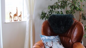 A brown leather armchair with a deer pillow, a window with white curtains, a plant, and a framed picture on the wall. - Stóri-Bakki Guesthouse with hot tub (Egilsstaðir)
