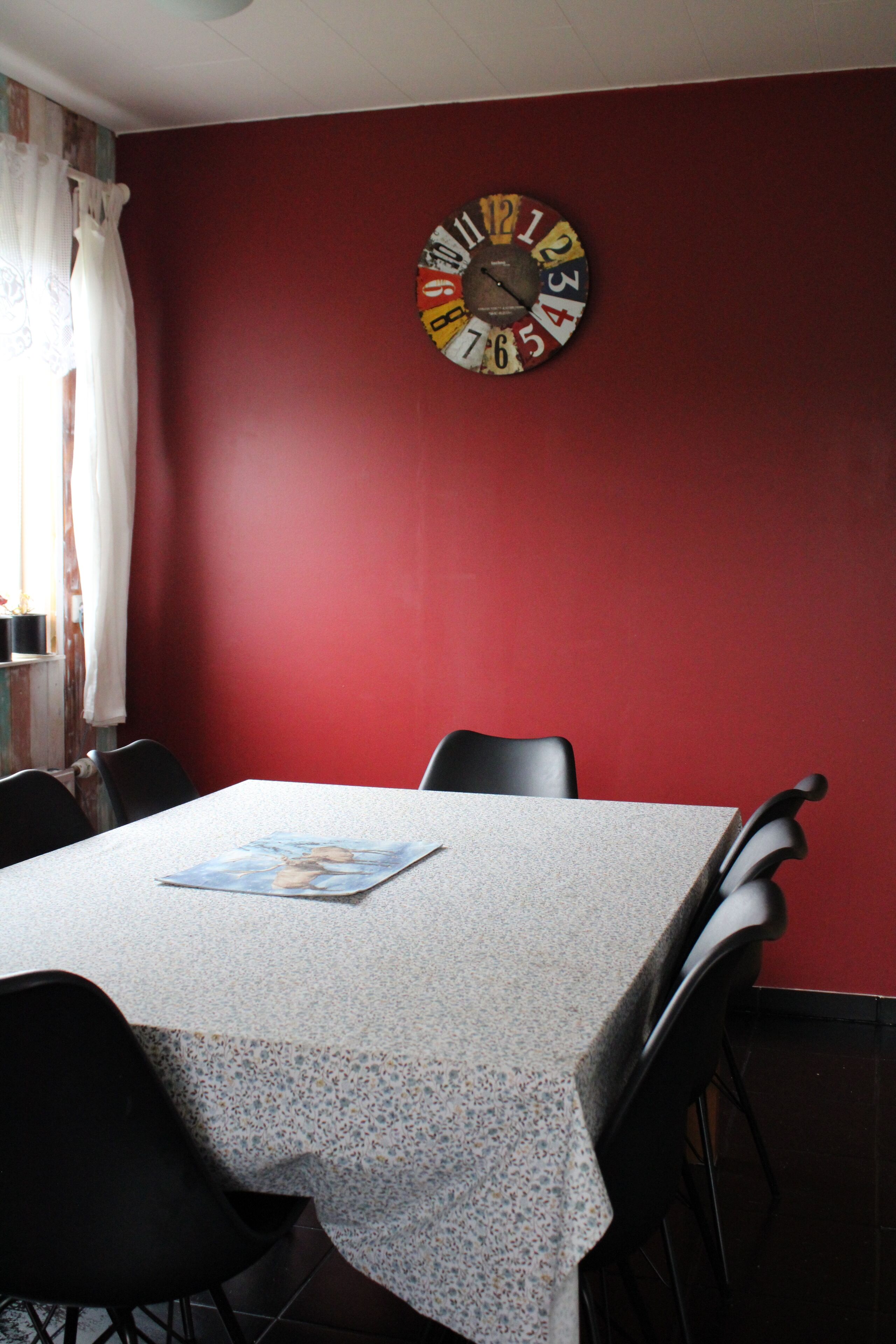 A dining area with a table set for four, chairs, a clock on the wall, and a window with curtains.
