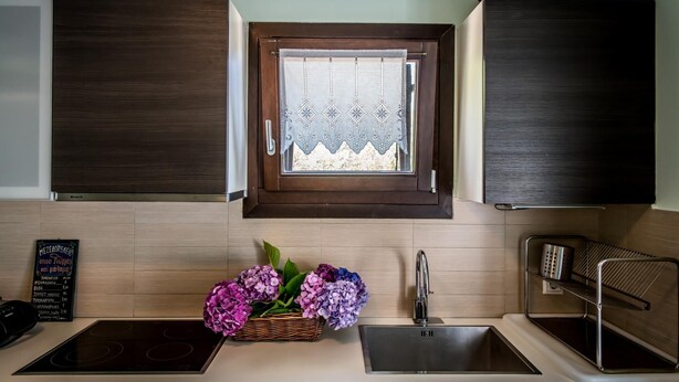 A kitchen with wooden cabinets, a window with lace curtains, a sink, and a basket of flowers. - Daphne House In Pelion (Zagora-Mouresi)
