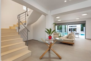 A modern interior with a staircase, a glass table with a red pot, a beige sofa, and a view of a swimming pool.
