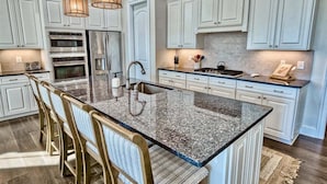 Une cuisine avec des meubles blancs, un îlot en granit, des tabourets de bar et des luminaires suspendus.