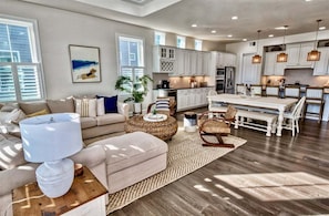 A spacious living room with a sofa, ottoman, and coffee table, a dining area with a table and chairs, and a kitchen with white cabinets and a refrigerator.